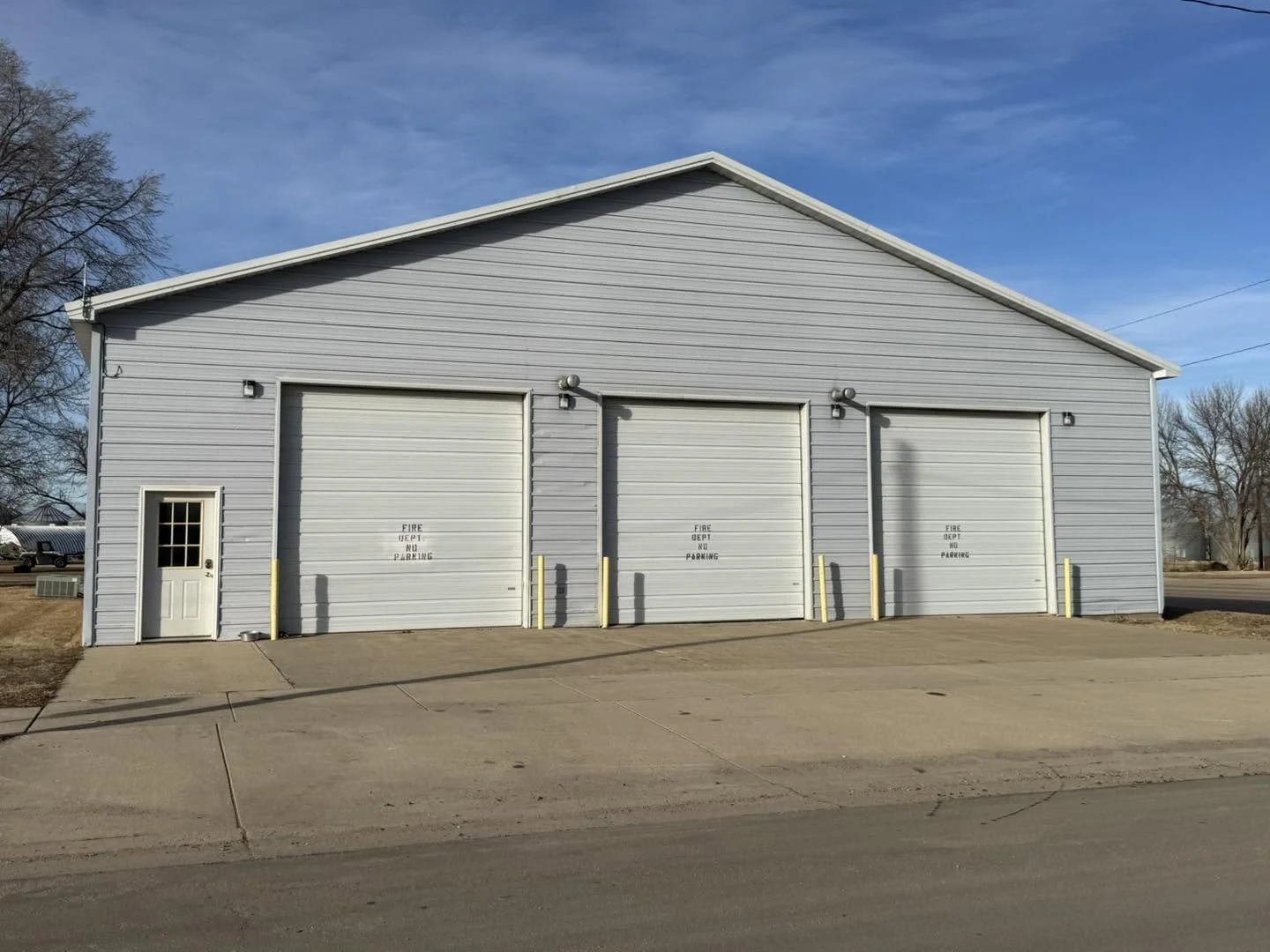A white industrial building with three garage doors and a small door on the left. The building is next to a concrete parking area, with a few trees and a clear blue sky in the background.