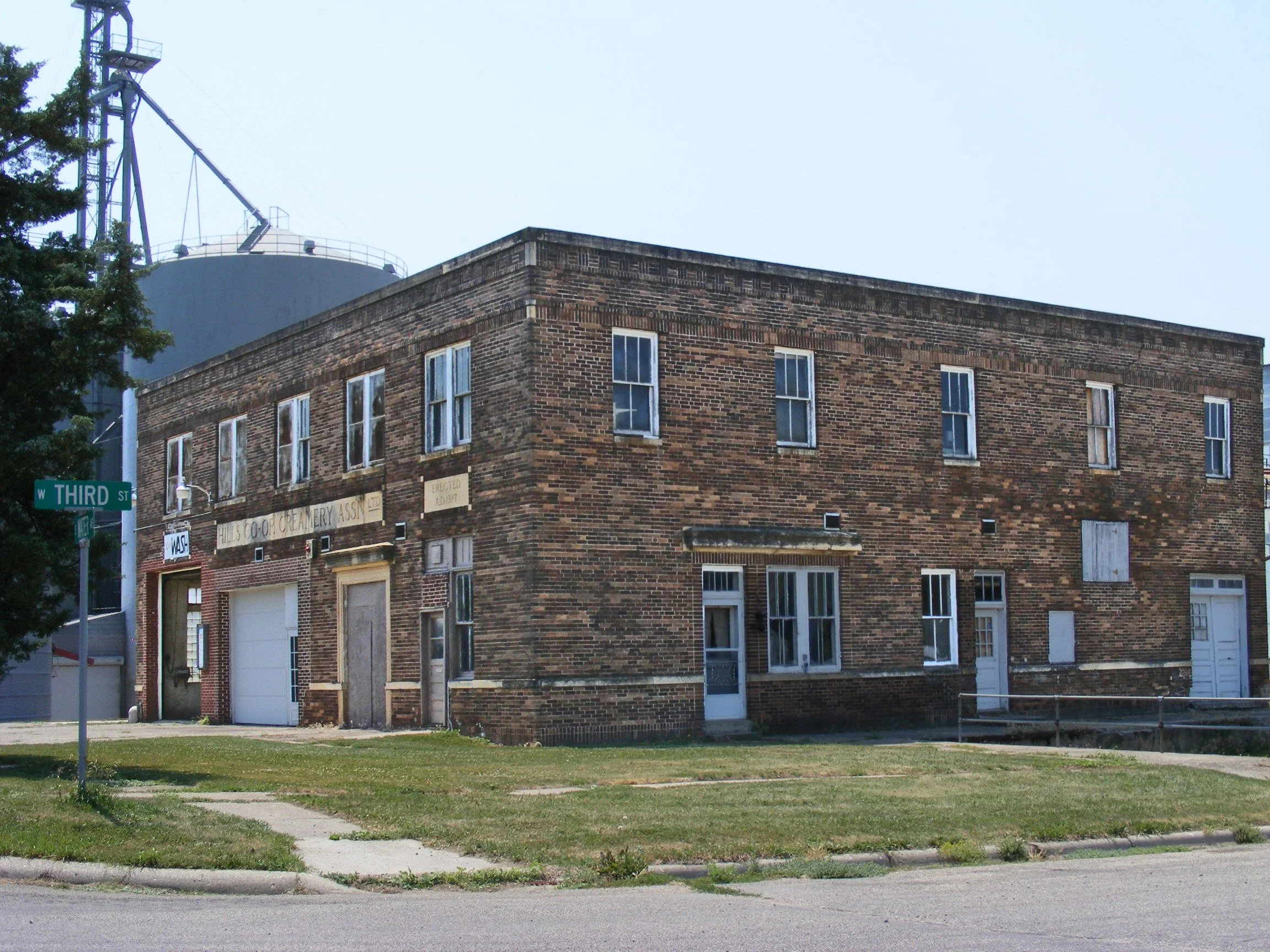 An intersection sign indicates West Third Street near a brick building with large windows. The building has old signage including a faded sign for a dairy association and another for a creamery. There is a grassy area and sidewalk in front of the building, along with a street curb.