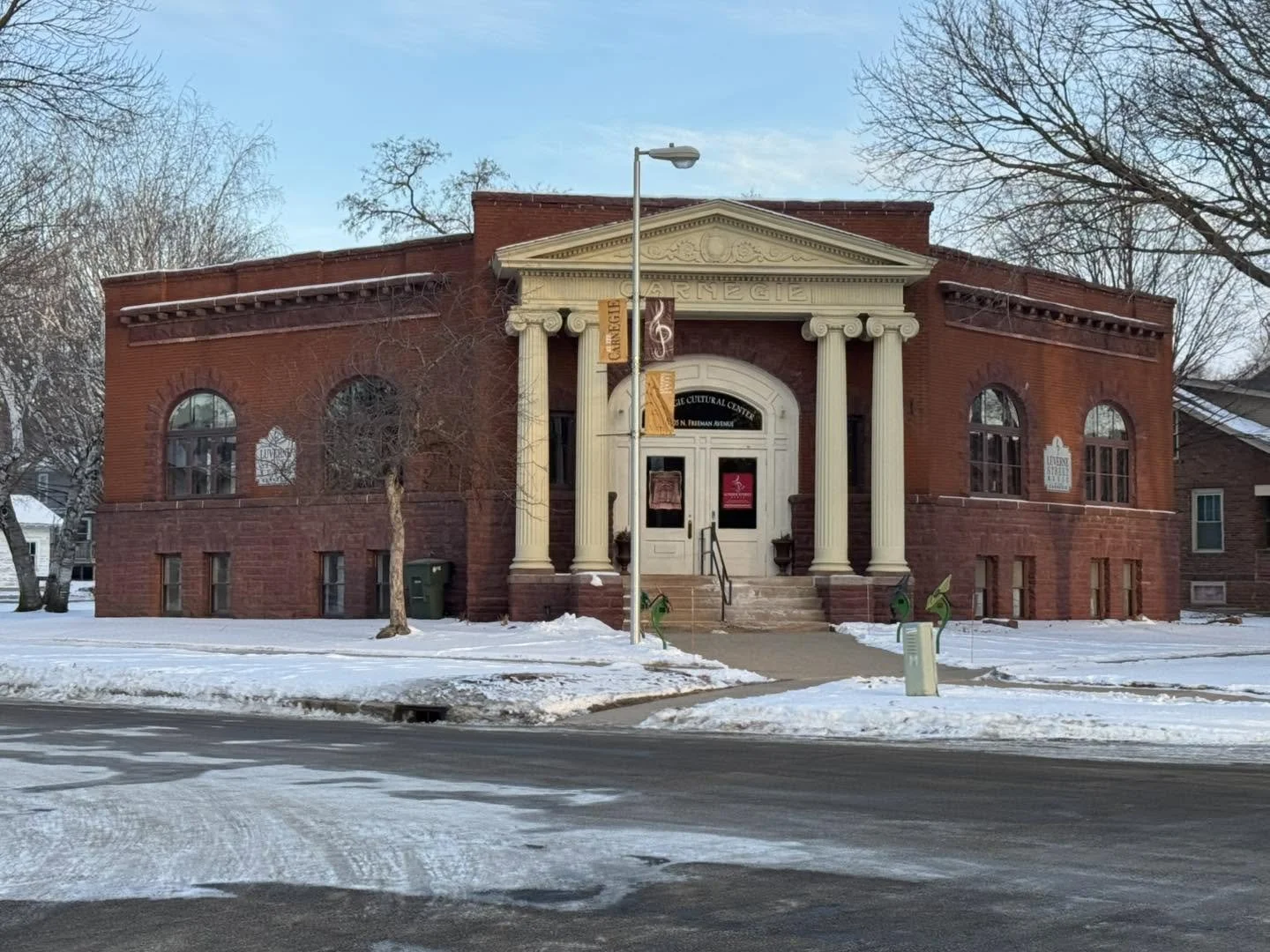 A red brick building with classical columns and a triangular pediment, labeled as a cultural center, is surrounded by snow on the ground and leafless trees.