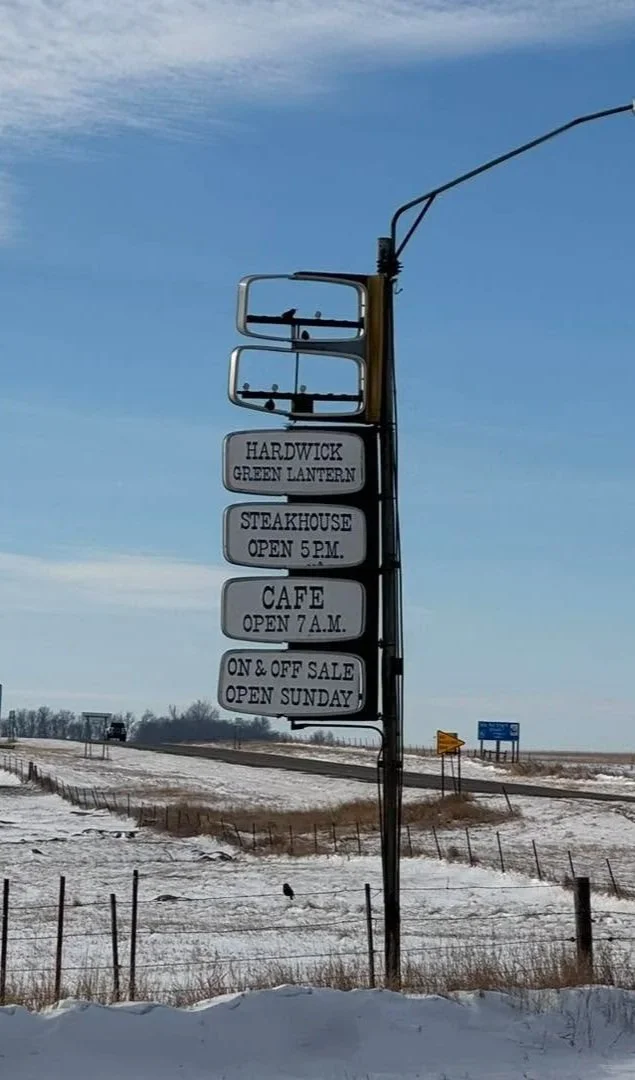 A tall roadside signpost with multiple signs showing local business names and hours, situated in a snowy rural area with a blue sky and a highway in the background.