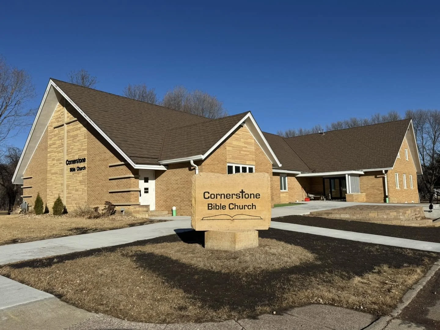 Exterior view of Cornerstone Bible Church, a brick building with a gabled roof and white trim. There is a large stone sign in front with the church's name, surrounded by a walkway and some landscaping.