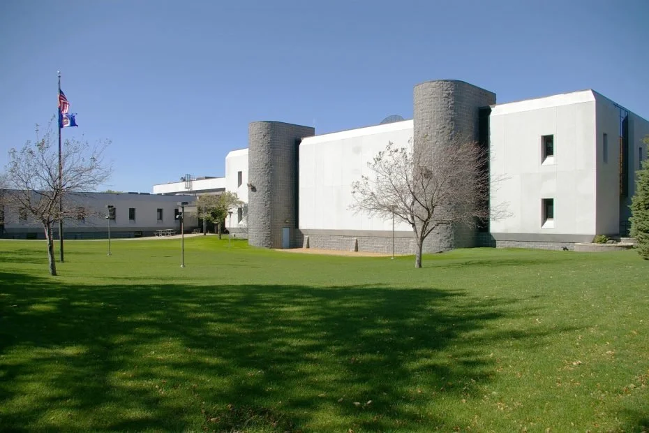 A modern white building with cylindrical gray stone towers, surrounded by a green lawn with a few trees and an American flag on a flagpole, under a clear blue sky.