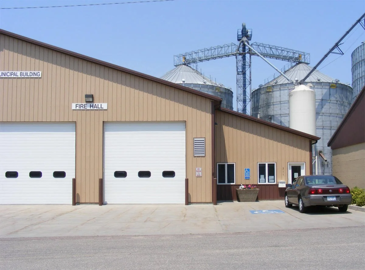 A fire hall building with two large garage doors, a sign labeled 'Municipal Building,' and a car parked in a designated disabled parking space outside. In the background, there are grain silos and a grain elevator structure.