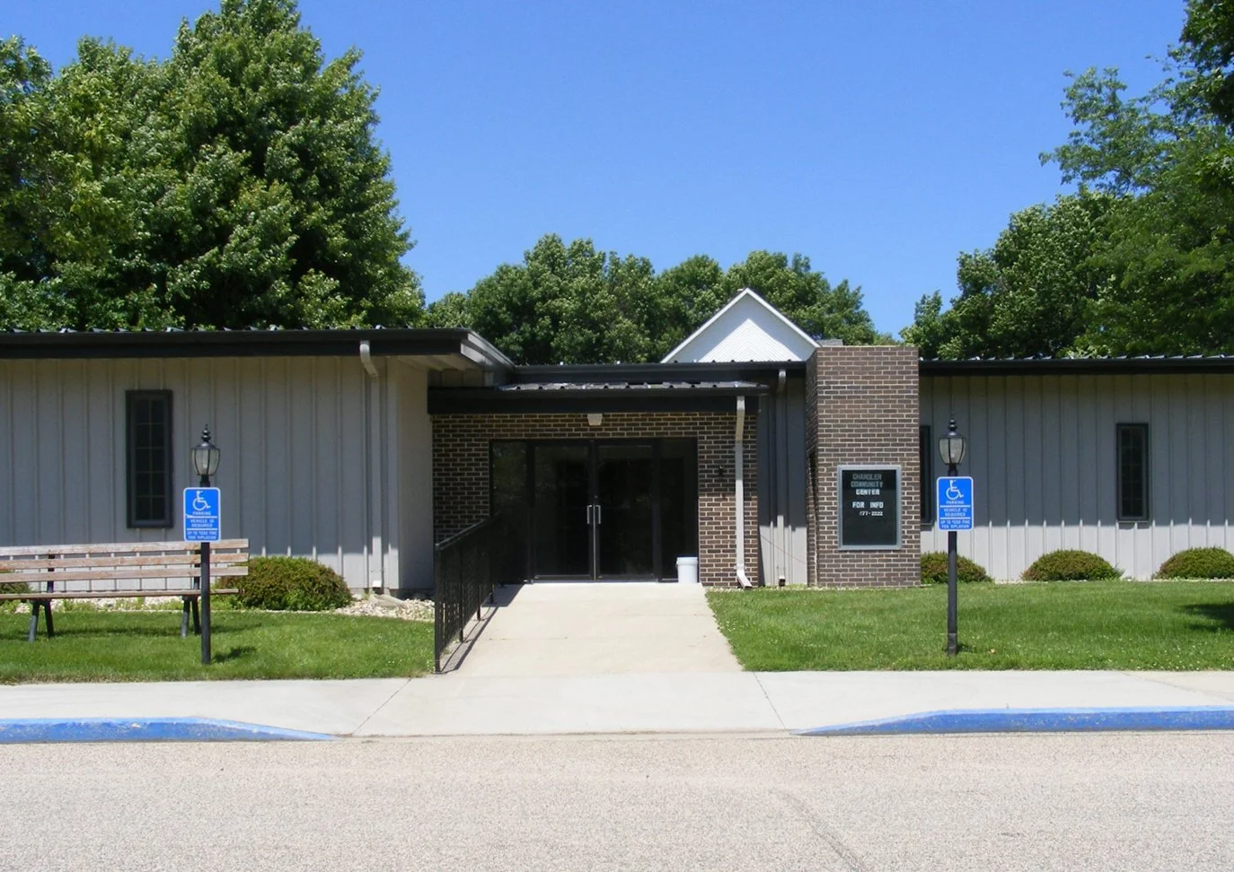 Chandler Community Center building with handicapped parking signs and benches in front, surrounded by trees on a sunny day.