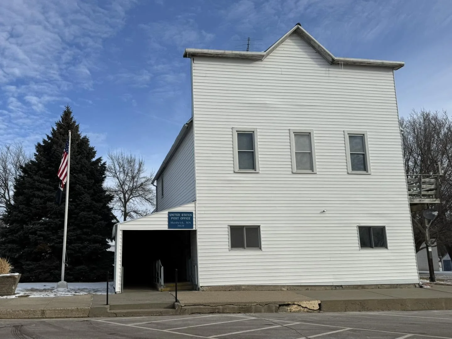 A white, multi-story building with a covered entrance labeled as a United States Post Office, located in Hardybeck, Minnesota. There is an American flag on a pole in front, and a large pine tree to the left of the building. The sky is partly cloudy, and some snow is visible on the ground.