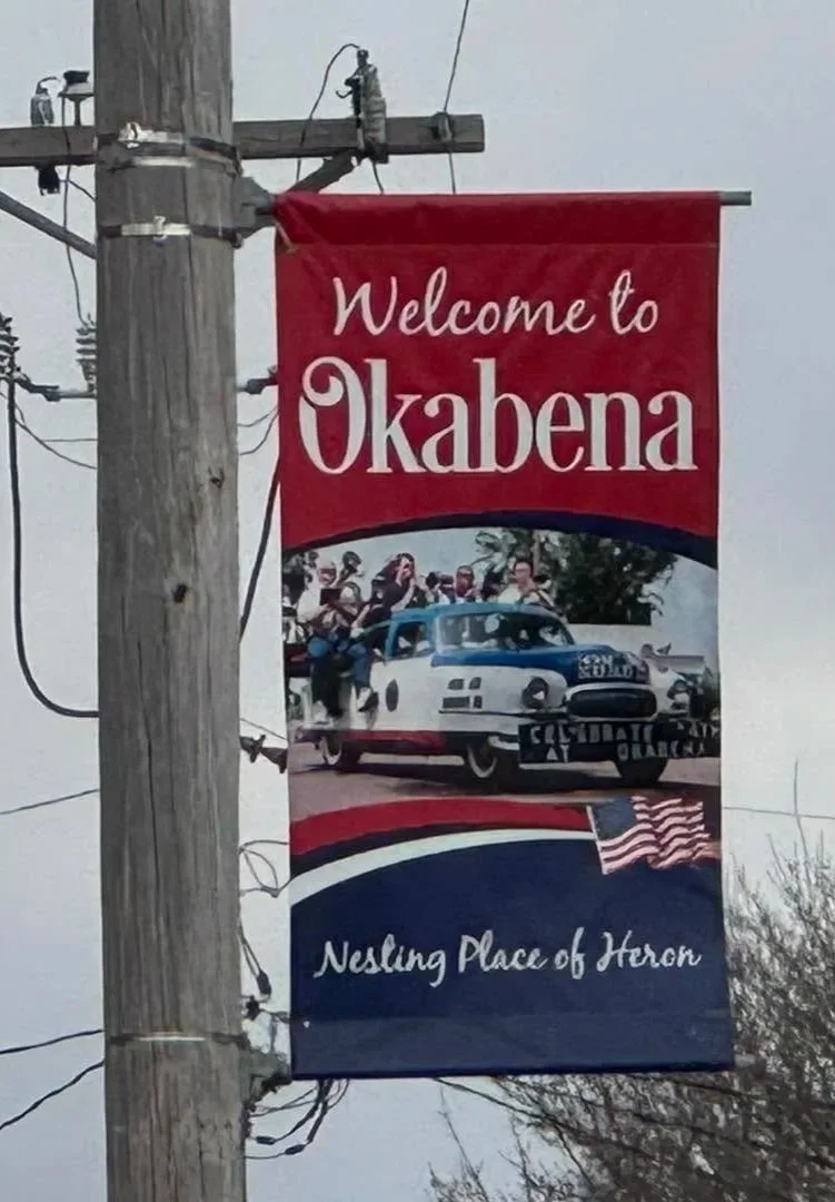 A vertical banner hanging from a utility pole with the text "Welcome to Okabena" and "Nesting Place of Horon." The banner features a vintage blue and white police car with a group of people, and small American flags at the bottom.
