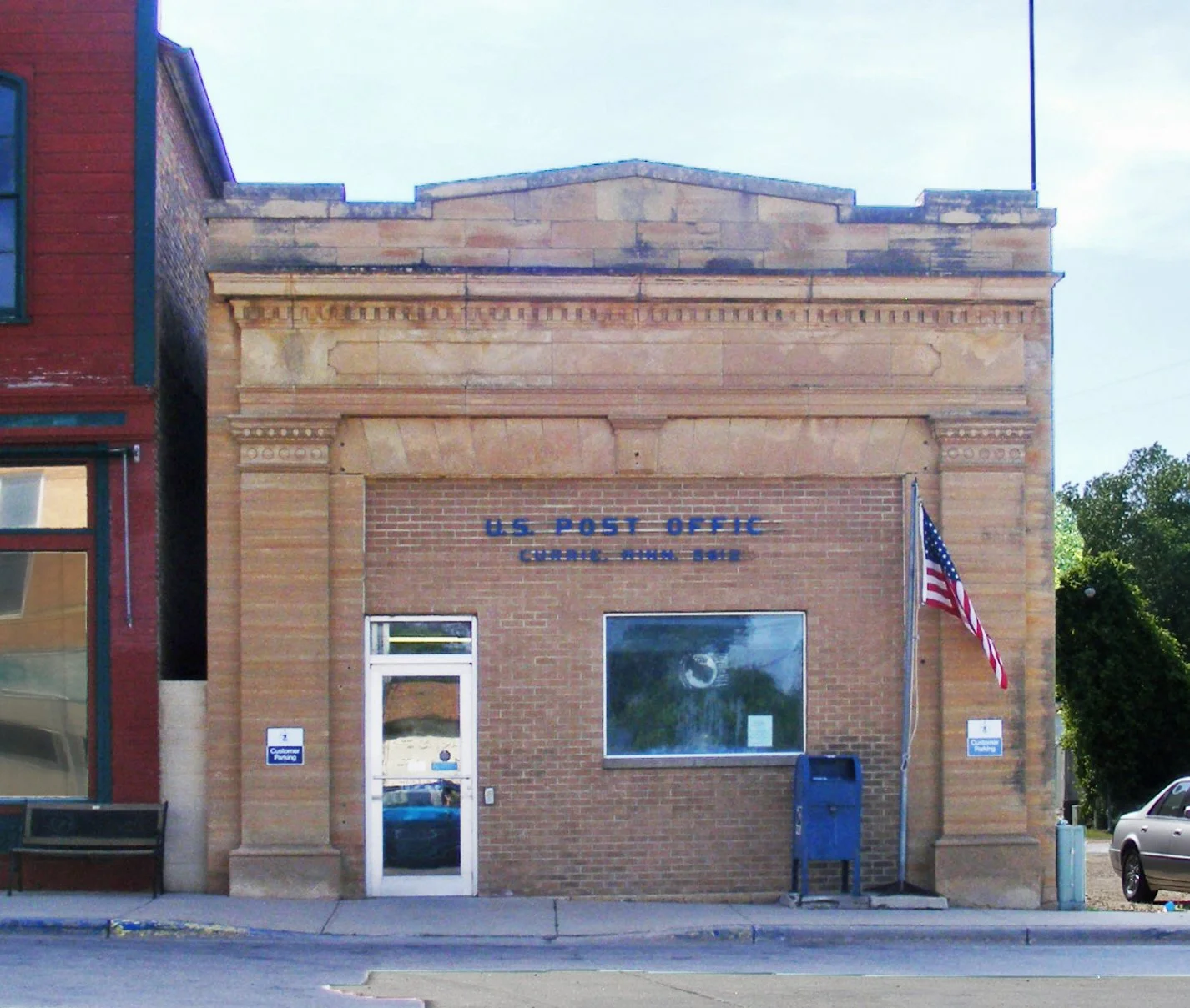 Front of a small brick building with a sign that reads 'U.S. Post Office, Garland, Iowa 52544', an American flag on a flagpole, a window, a door, and a mailbox outside.