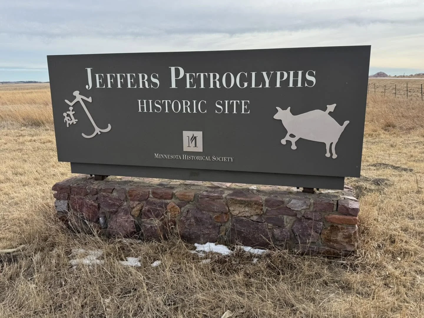 Sign for Jeffers Petroglyphs Historic Site in Minnesota, with an anchor symbol and a cow silhouette, set on a stone base in an open field.