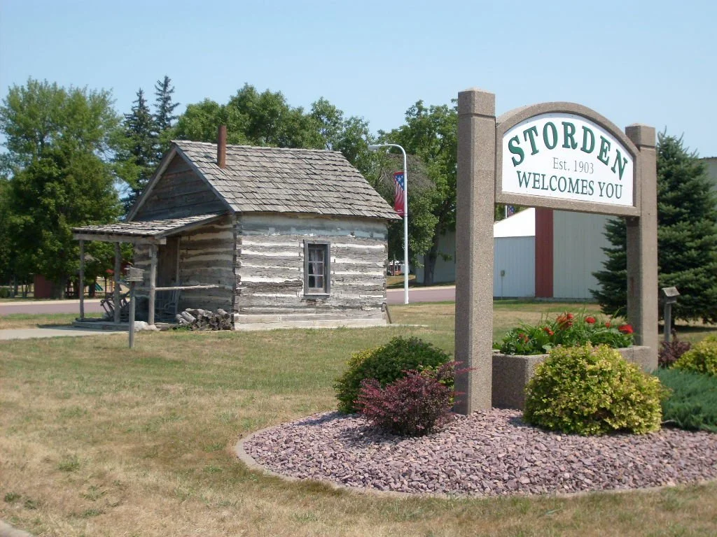 A wooden historic log cabin with a covered porch and a small chimney, located behind a landscaped area with shrubs, next to a sign that reads "Storden Est. 1903 Welcomes You" and surrounded by trees.