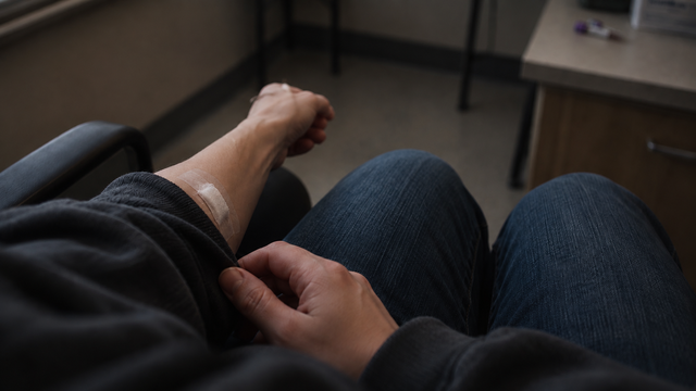 Person sitting in a chair with a bandage on their left arm, holding their hand.