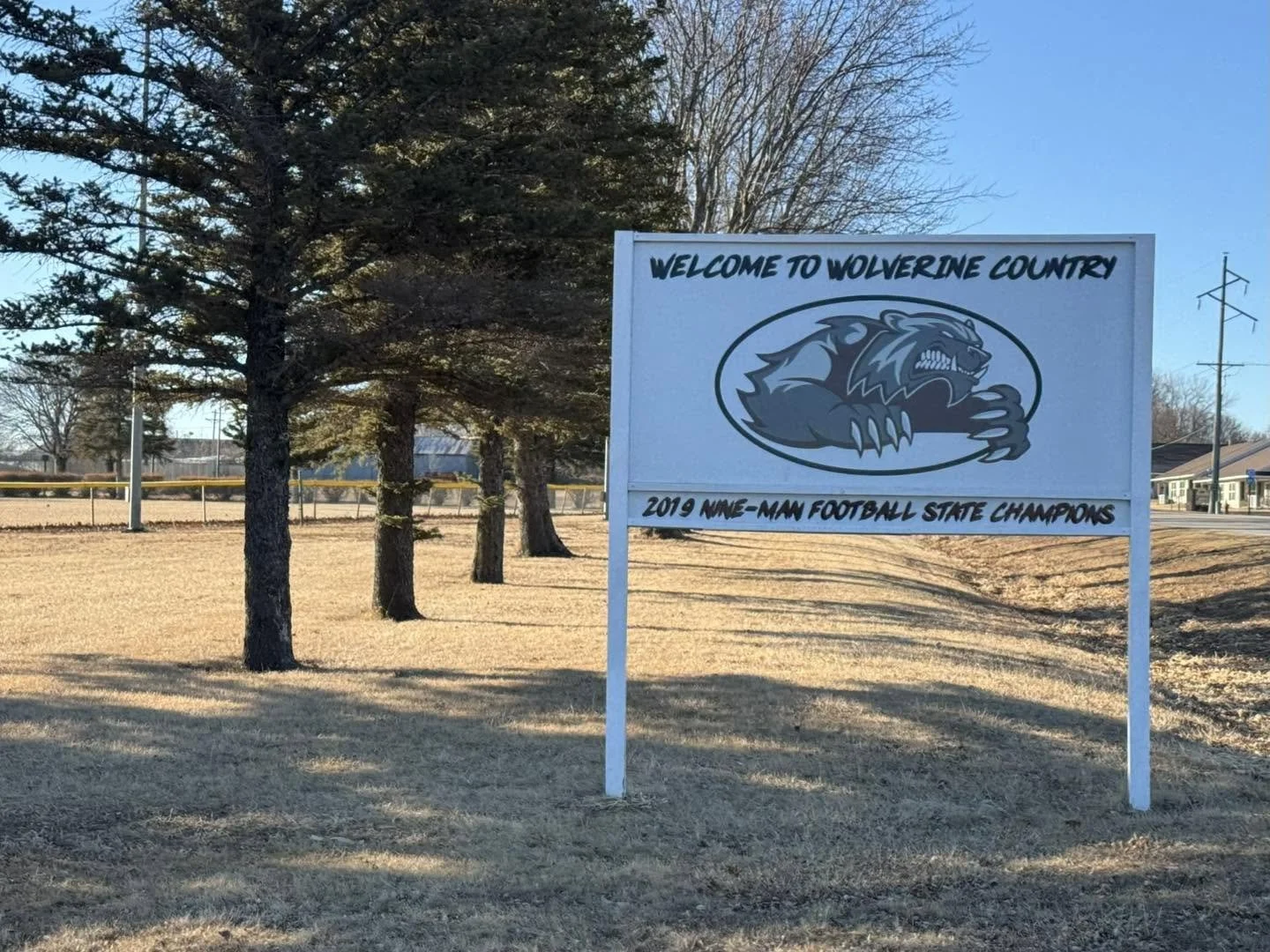 A sign welcoming visitors to Wolverine County, featuring a large wolf mascot, with trees, grass, and a few buildings in the background under a clear blue sky.