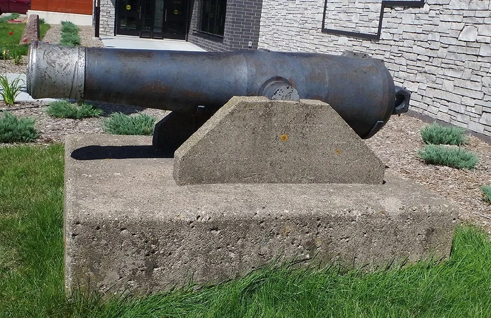 An old, large cannon mounted on a concrete base outdoors, with a stone building and landscaped garden in the background.