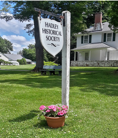 A sign for the Hadley Historical Society hanging from a wooden post in a grassy park area with a flower pot of pink flowers at its base, residential houses and trees in the background.