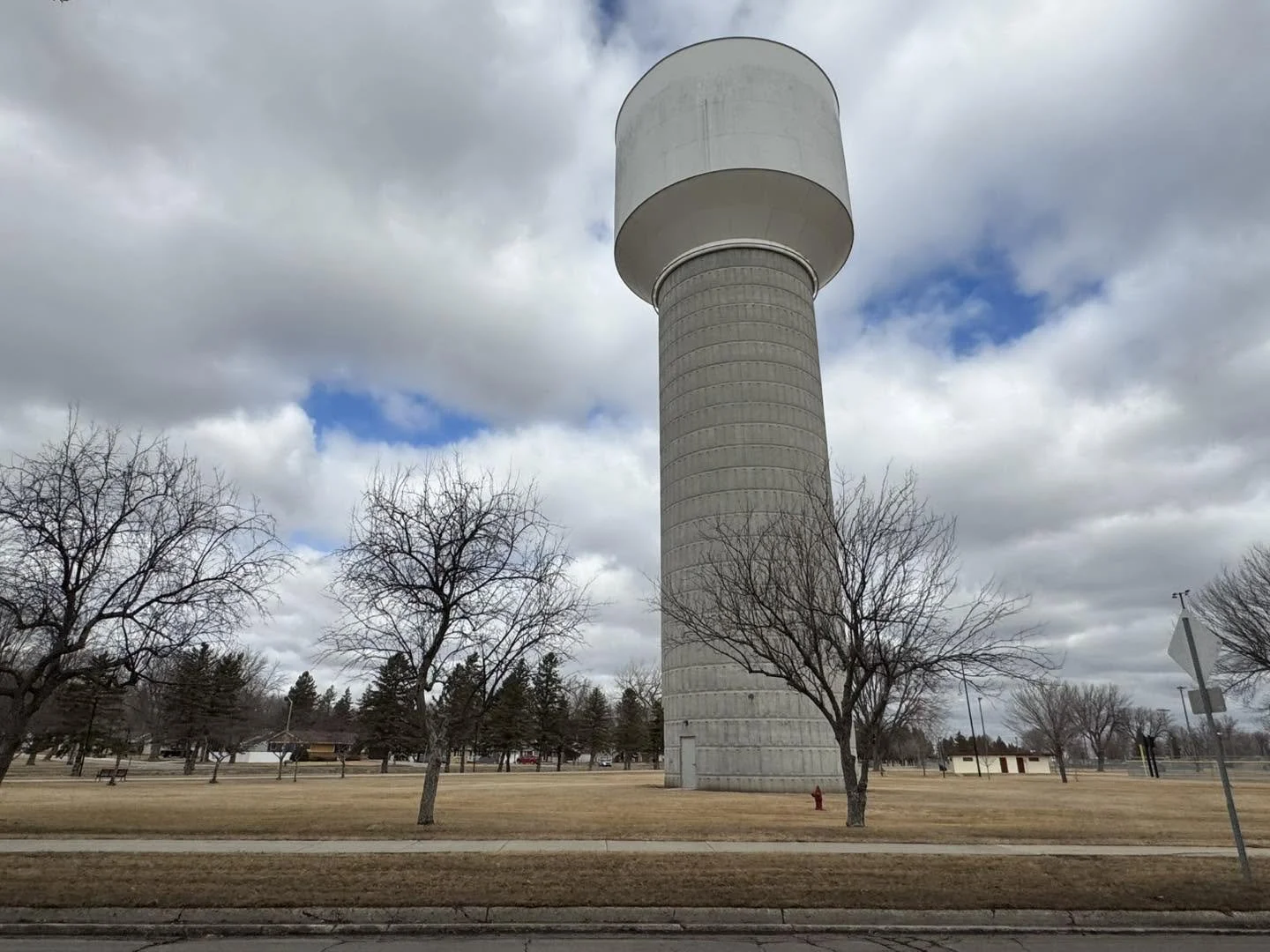 A tall, cylindrical water tower with a large, rounded top, located in a park with leafless trees and a cloudy sky overhead.