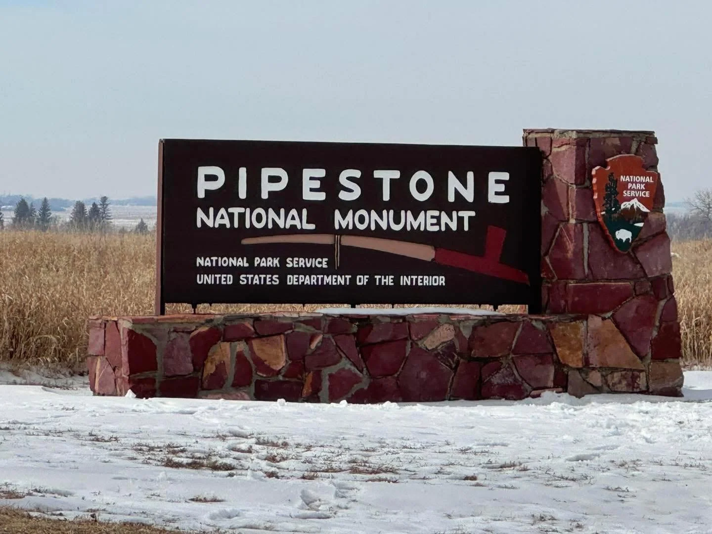 Signs at the entrance of Pipestone National Monument, a designated site for the National Park Service in the United States, with snow on the ground and a field in the background.