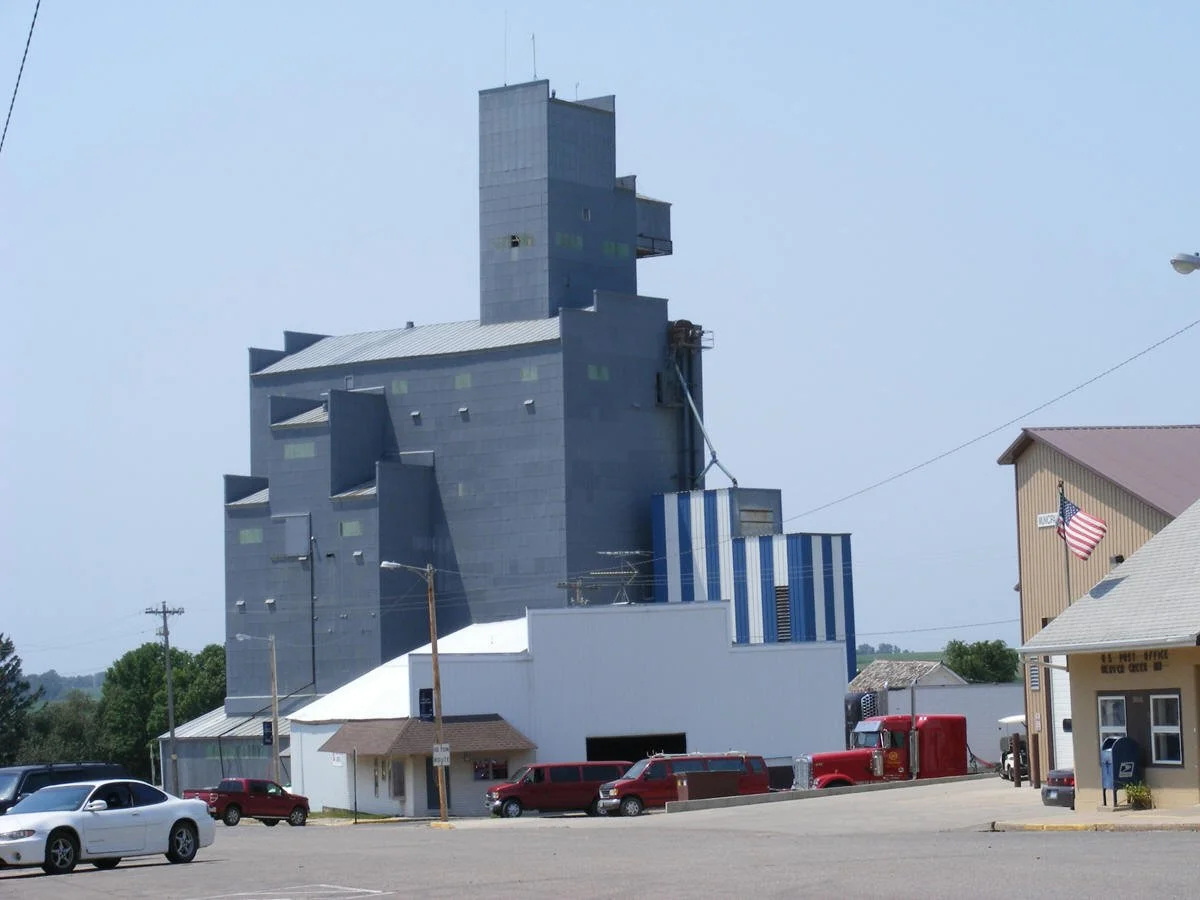 A small town scene with a large, modern, multi-level gray building and a nearby beige building flying an American flag. Several cars are parked in the foreground, and power lines are visible. The sky is clear and blue.