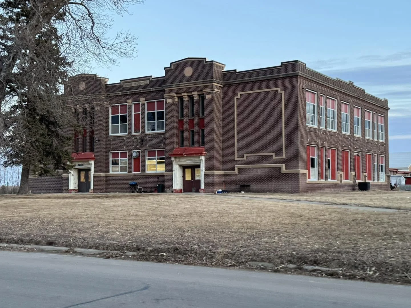 A large, old brick school building with multiple windows and red accents, surrounded by a barren lawn and a sidewalk in front.