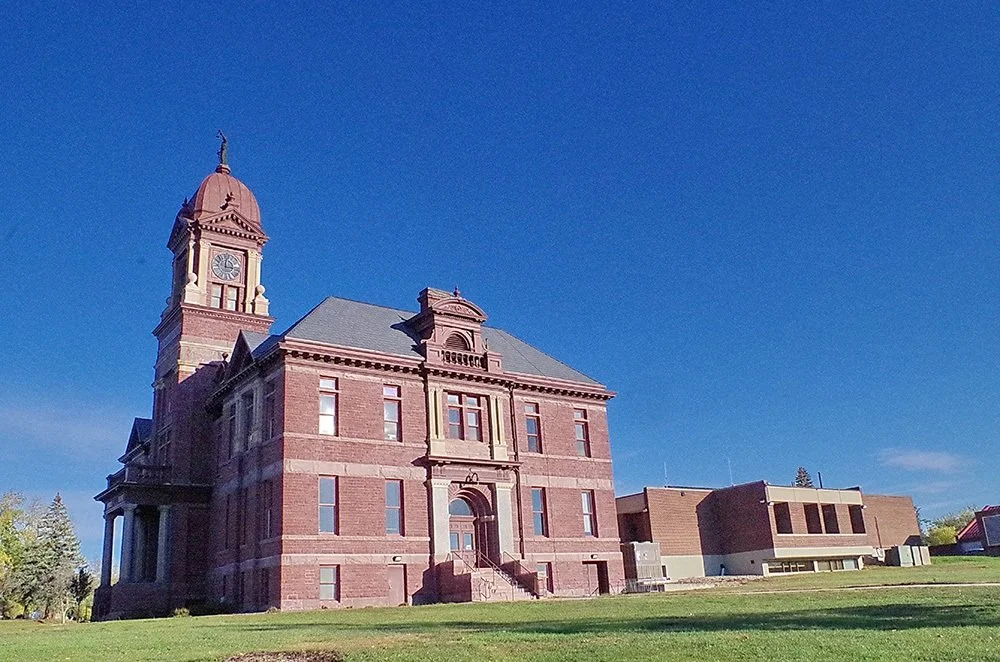 A historic brick courthouse with a clock tower and a gray roof, set against a clear blue sky, with a lawn in the foreground.