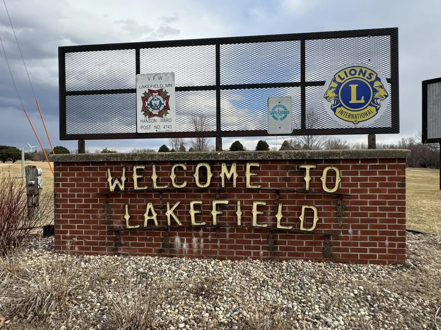 A welcome sign for Lakefield, Minnesota, with a brick base and metal letters, and a large billboard above displaying logos for veterans and Lions Club International.