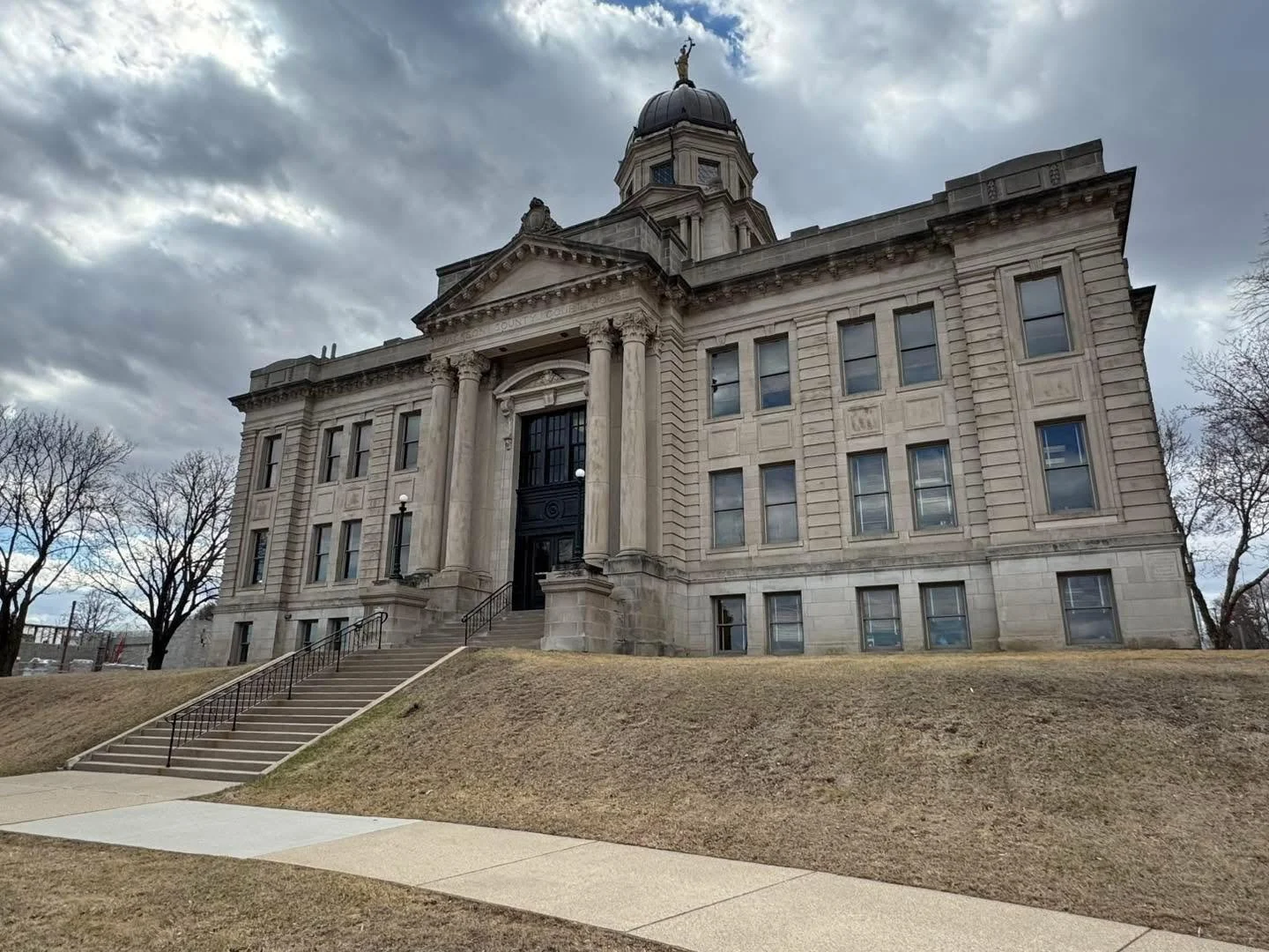 A historic government building with a domed roof topped with a statue, stone steps leading to a dark entrance, and a grassy area in the foreground under a cloudy sky.
