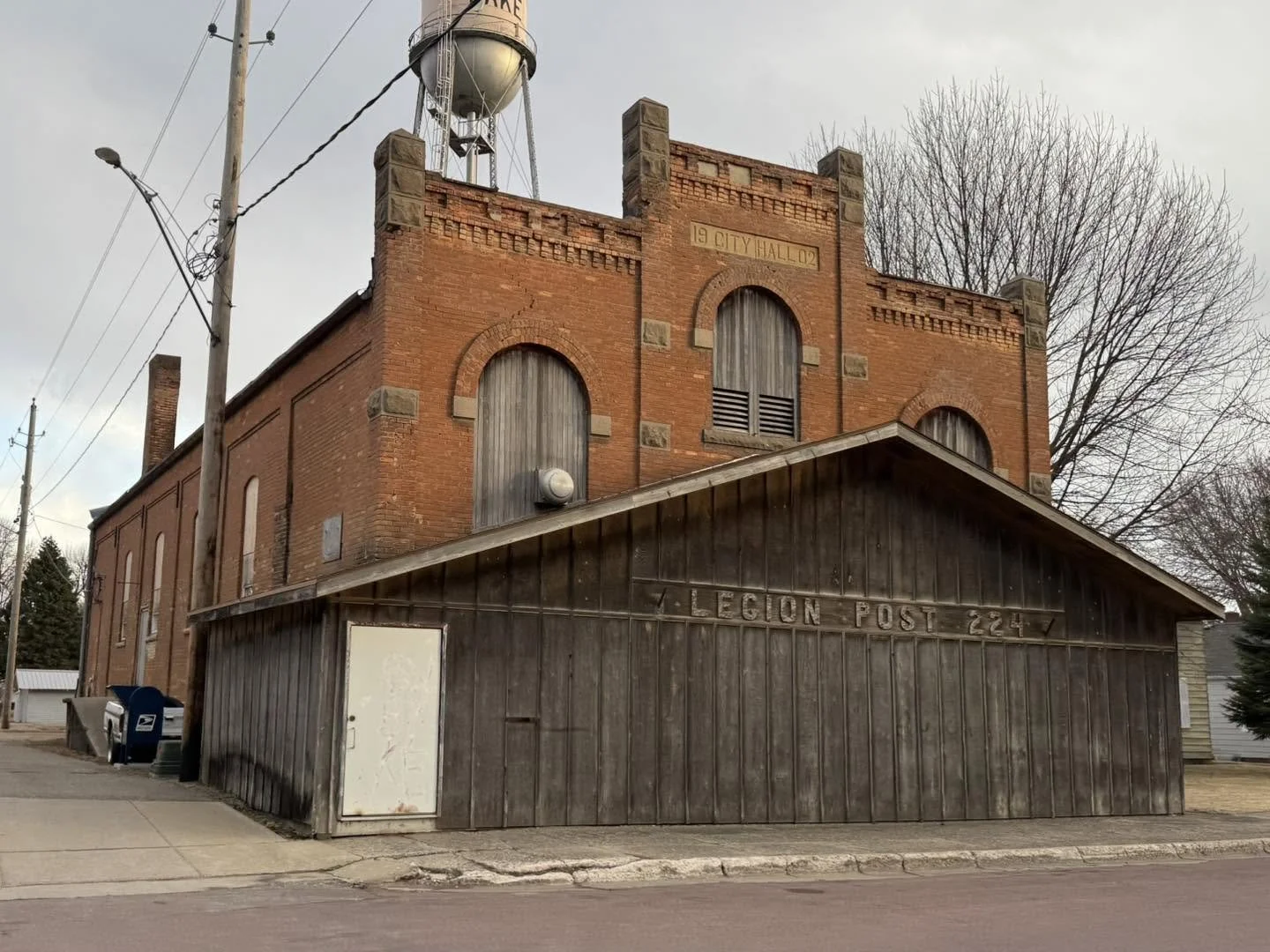 A brick building with a wooden addition labeled 'Legion Post 224' and a water tower behind it, located in a small town.