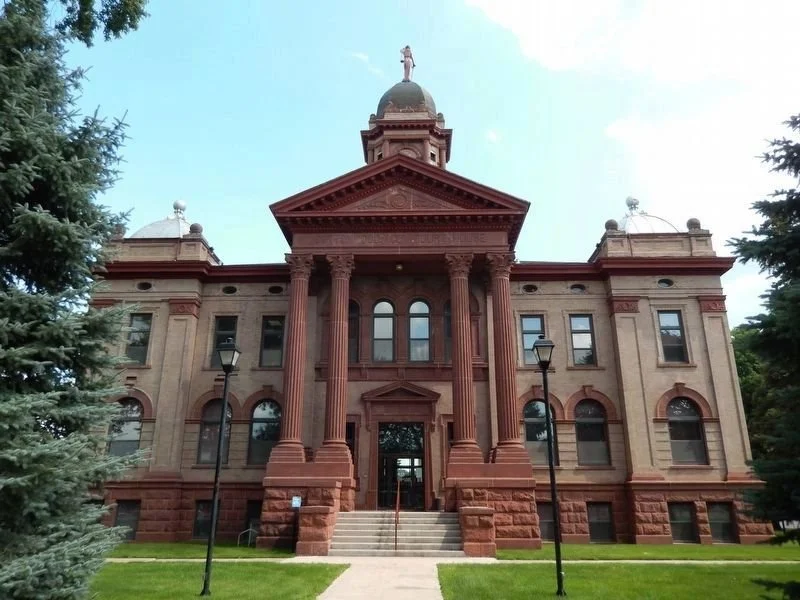 Front view of a historic courthouse with red brick and stone facade, large columns, and a central dome with a statue on top, surrounded by green lawn and trees.