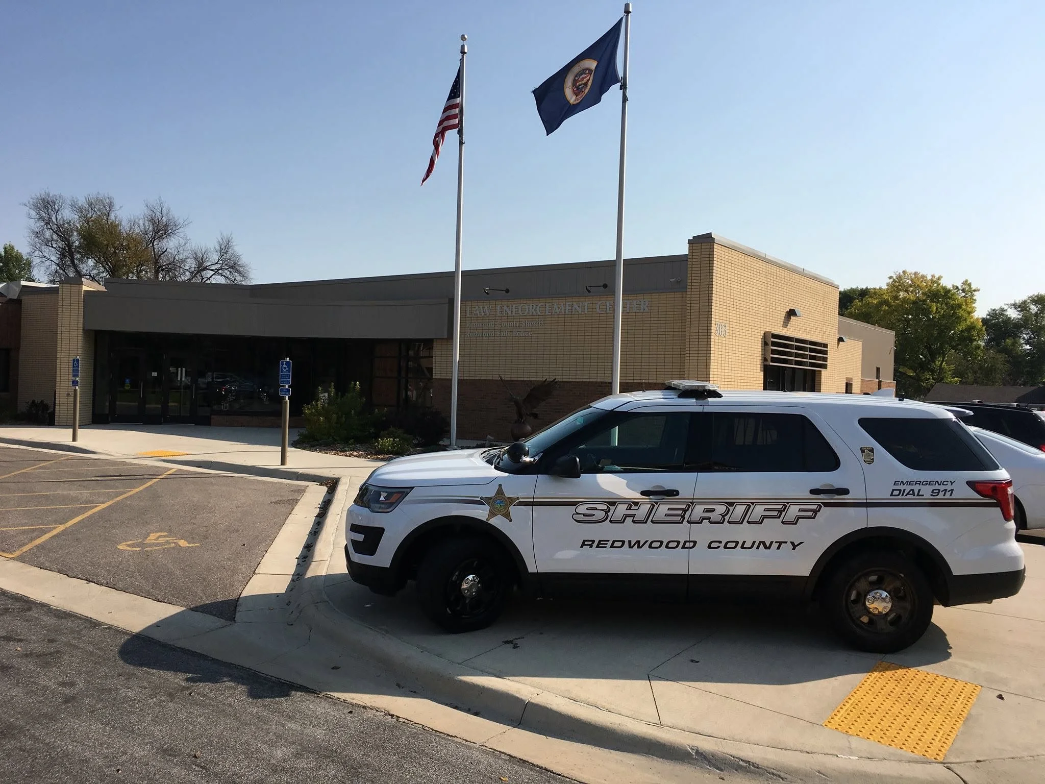 A police vehicle with 'Sheriff Redwood County' written on it is parked in front of a building labeled 'Law Enforcement Center.' Behind the vehicle, there is a flagpole with the American flag and another flag. The scene is outdoors with trees in the background, and there are parking spaces and handicapped parking signs visible.