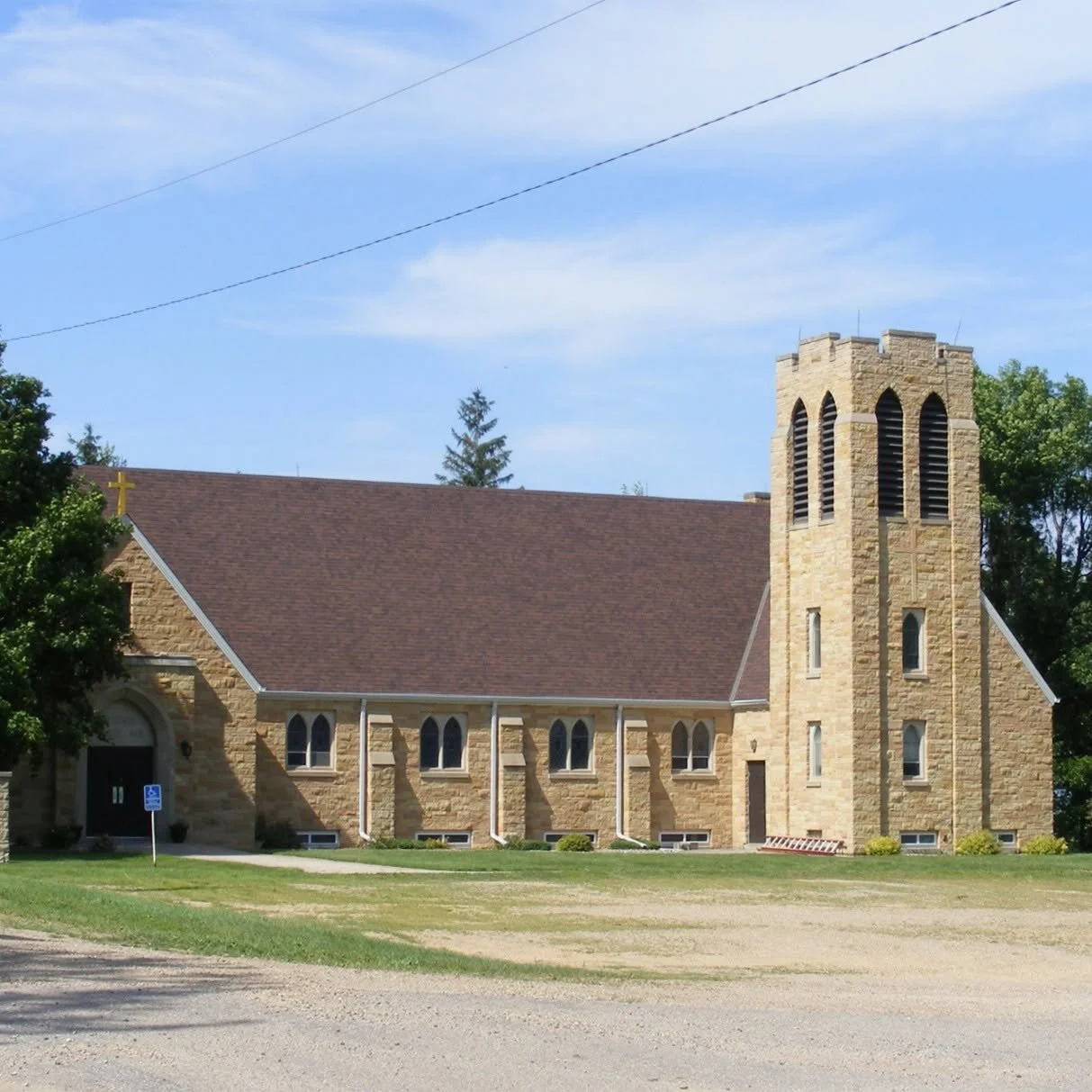 A beige stone church with a tall bell tower and arched windows, surrounded by trees and a grassy area under a blue sky.