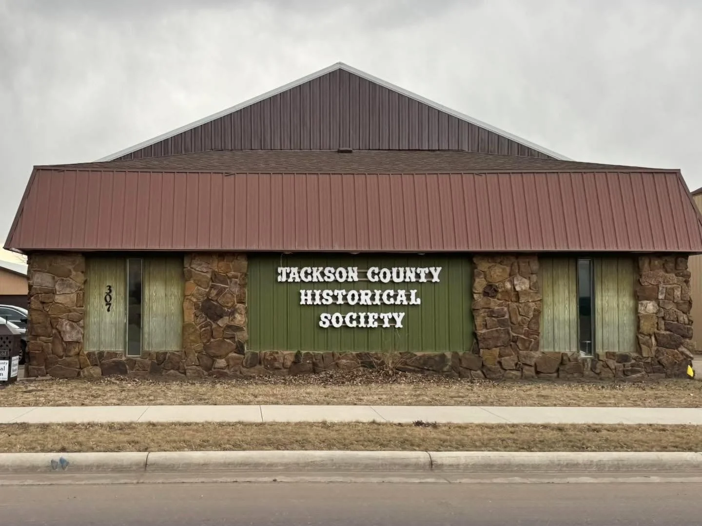 Front of a building with stone and green panel walls, red metal roof, and a sign that reads 'Tackson County Historical Society'.