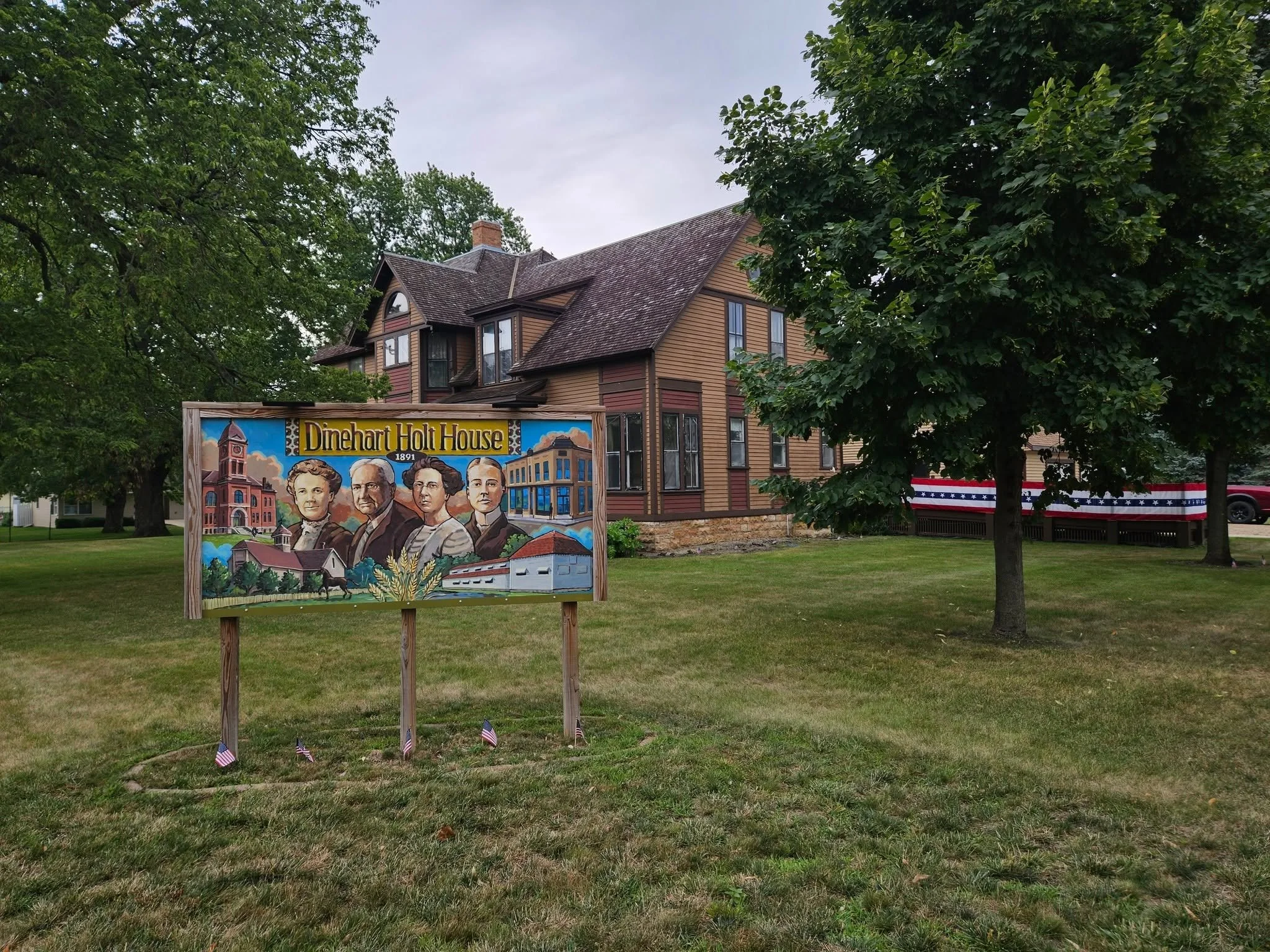 A colorful mural sign depicting historical figures and buildings from Dinehart Holt House, with trees and a house with a distinctive roof in the background.