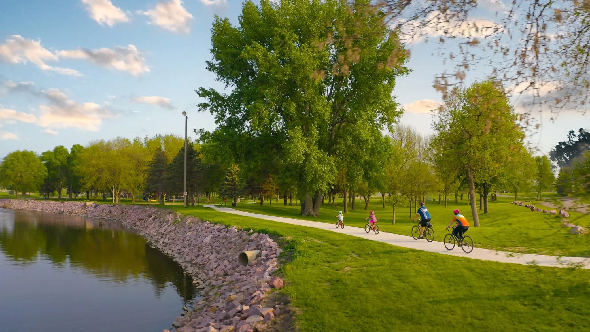 A park scene with a group of children and adults riding bicycles along a paved path next to a calm body of water. The park is filled with lush green trees, and the sky is partly cloudy.