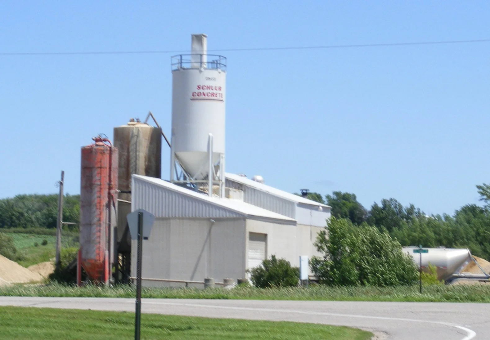 Rural grain silo and storage facility with surrounding grass and trees, under a clear blue sky.