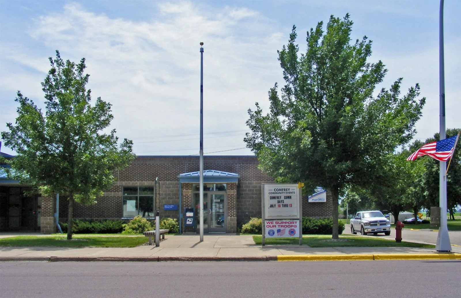 Post office building with trees, American flags, and a sidewalk in front. Sign advertising community events and support for troops.