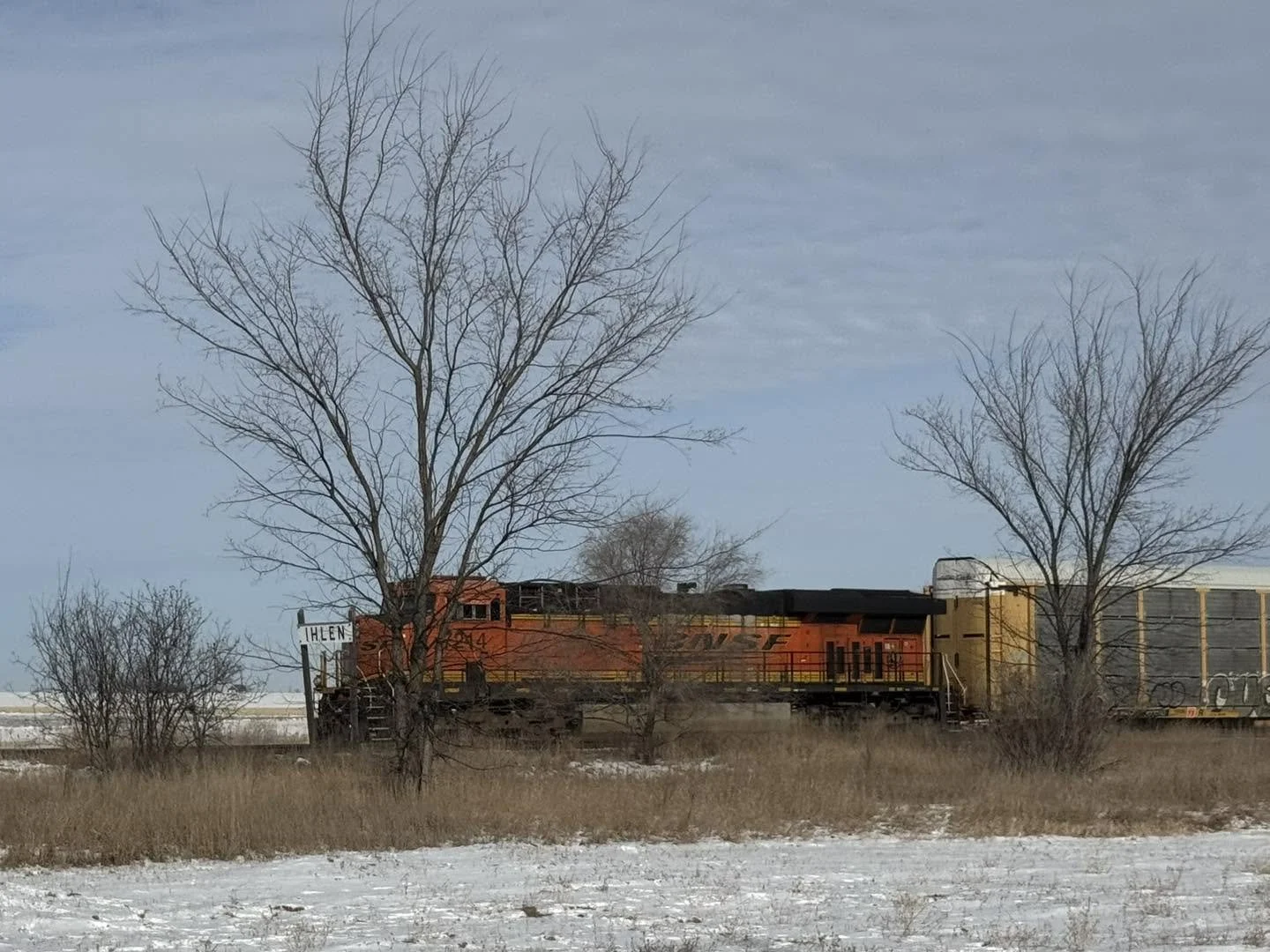 A red and yellow train moving through a winter landscape with bare trees and snow-covered ground, under a cloudy sky.