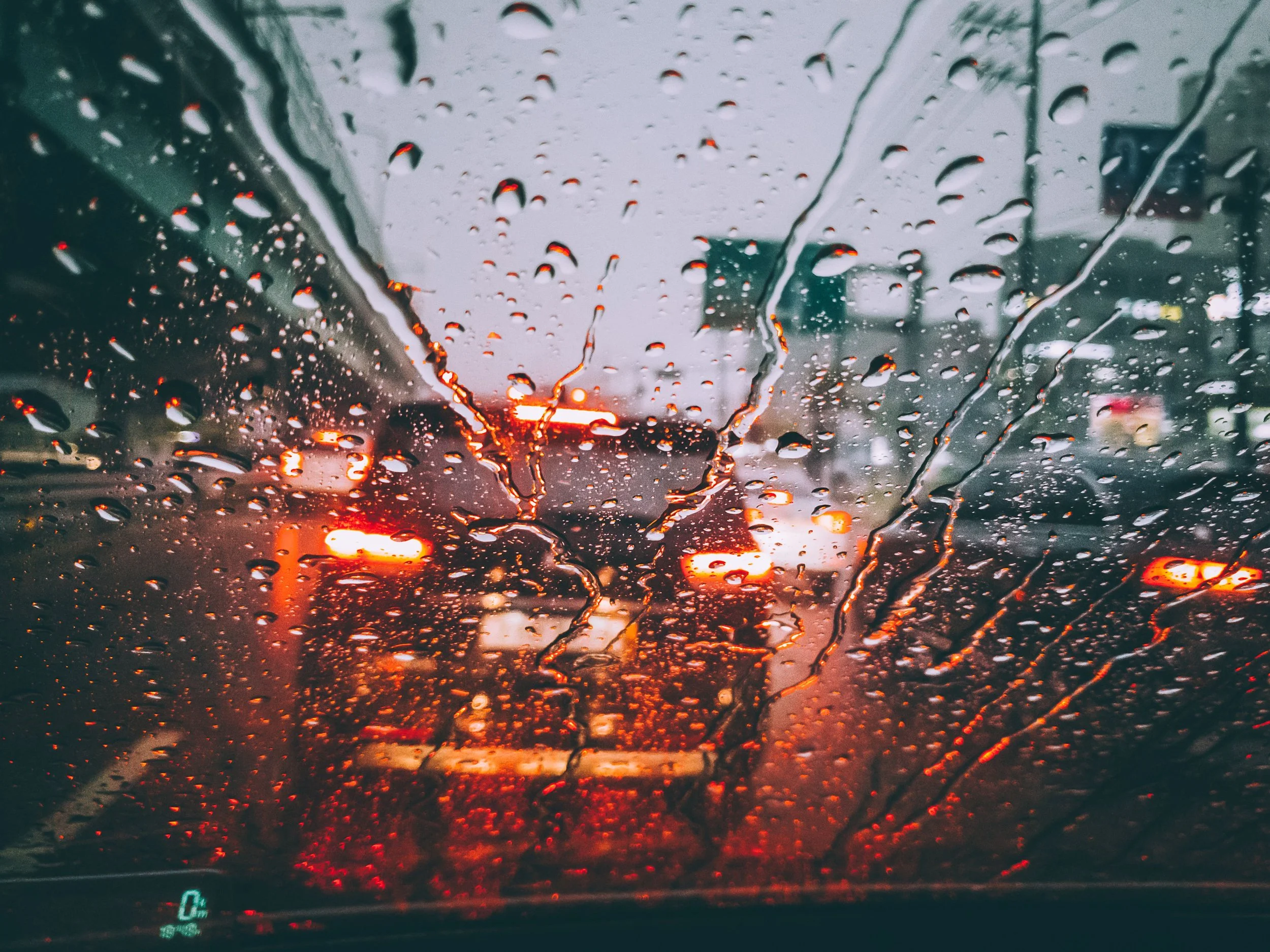 View through a car windshield on a rainy day, with water droplets on the glass and blurred red and orange taillights of cars ahead.