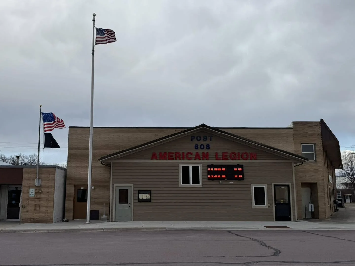 A building with a sign that reads 'Post 608 American Legion' and two flags on flagpoles in front, one American flag and one black flag. The sky is overcast.