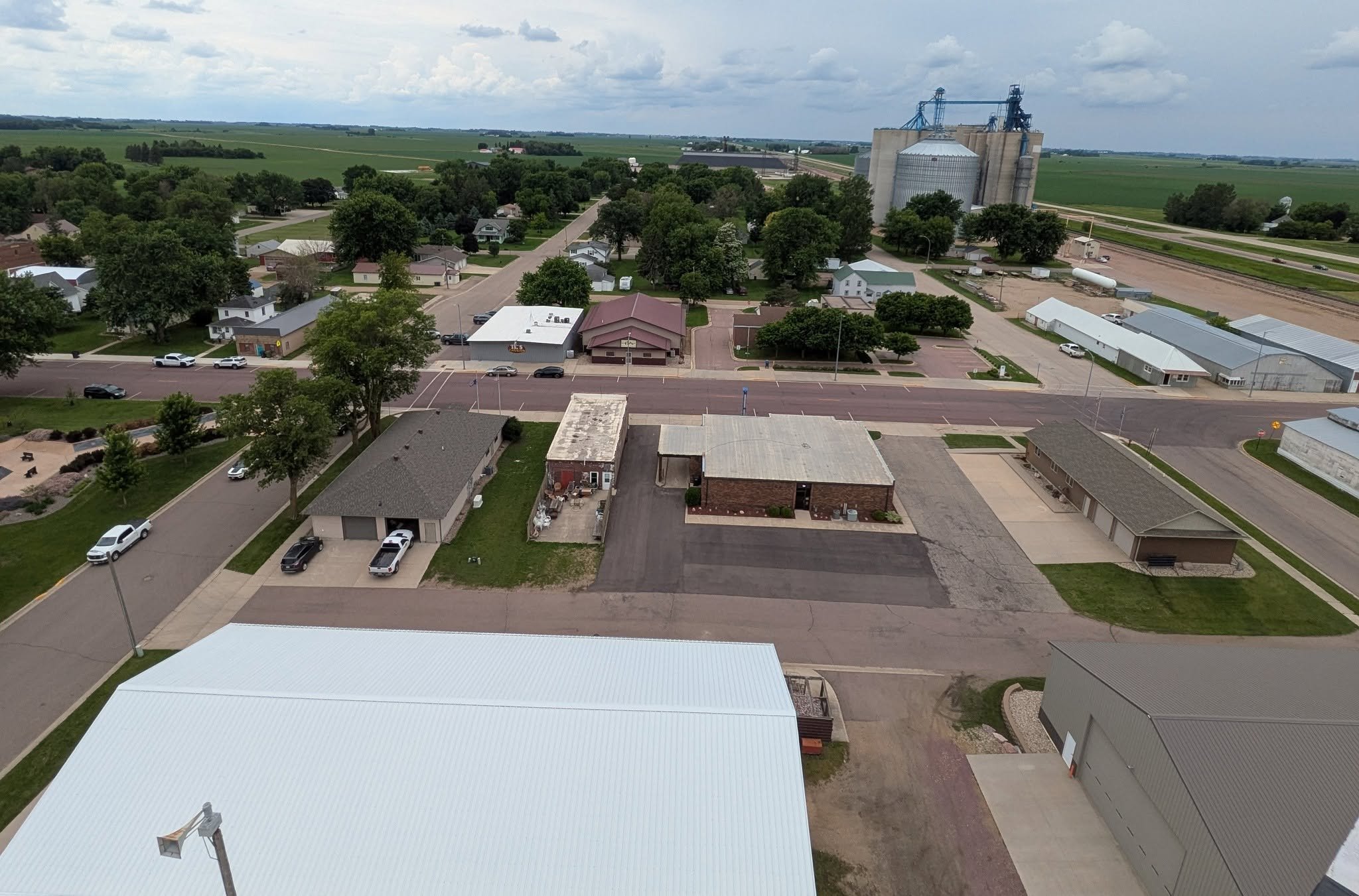Aerial view of a small town with residential houses, a commercial building, and a large industrial grain elevator in the distance, surrounded by farmland under cloudy skies.