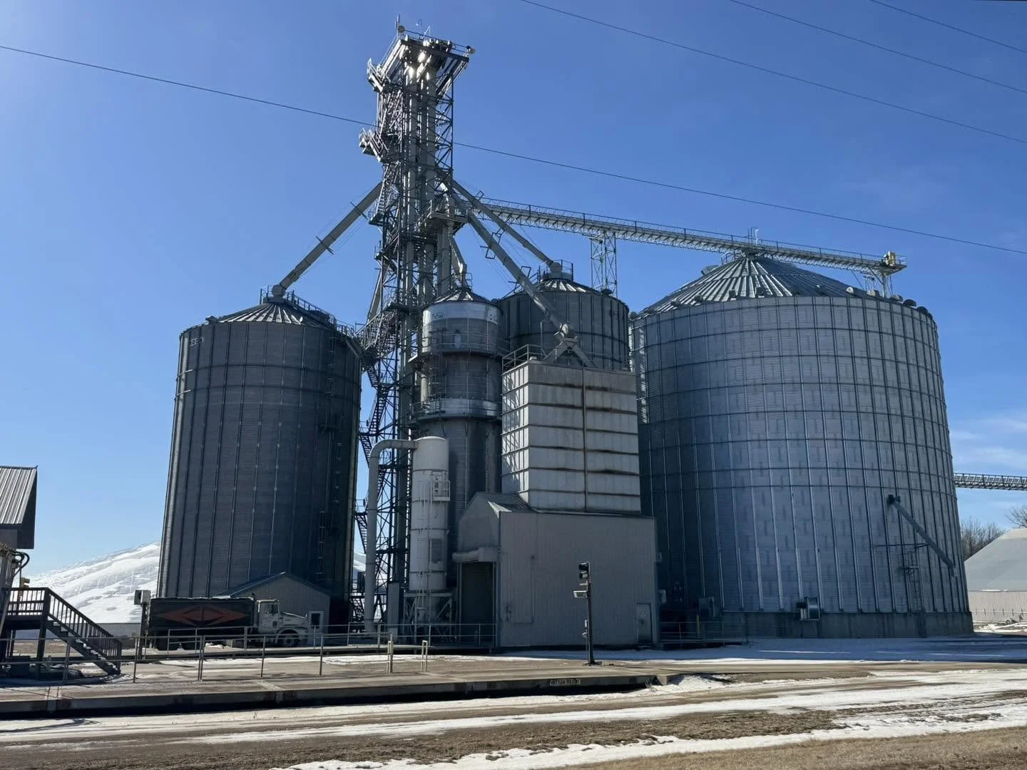 Large industrial grain silos with a conveyor system and machinery, set against a clear blue sky with snowy ground in the foreground.