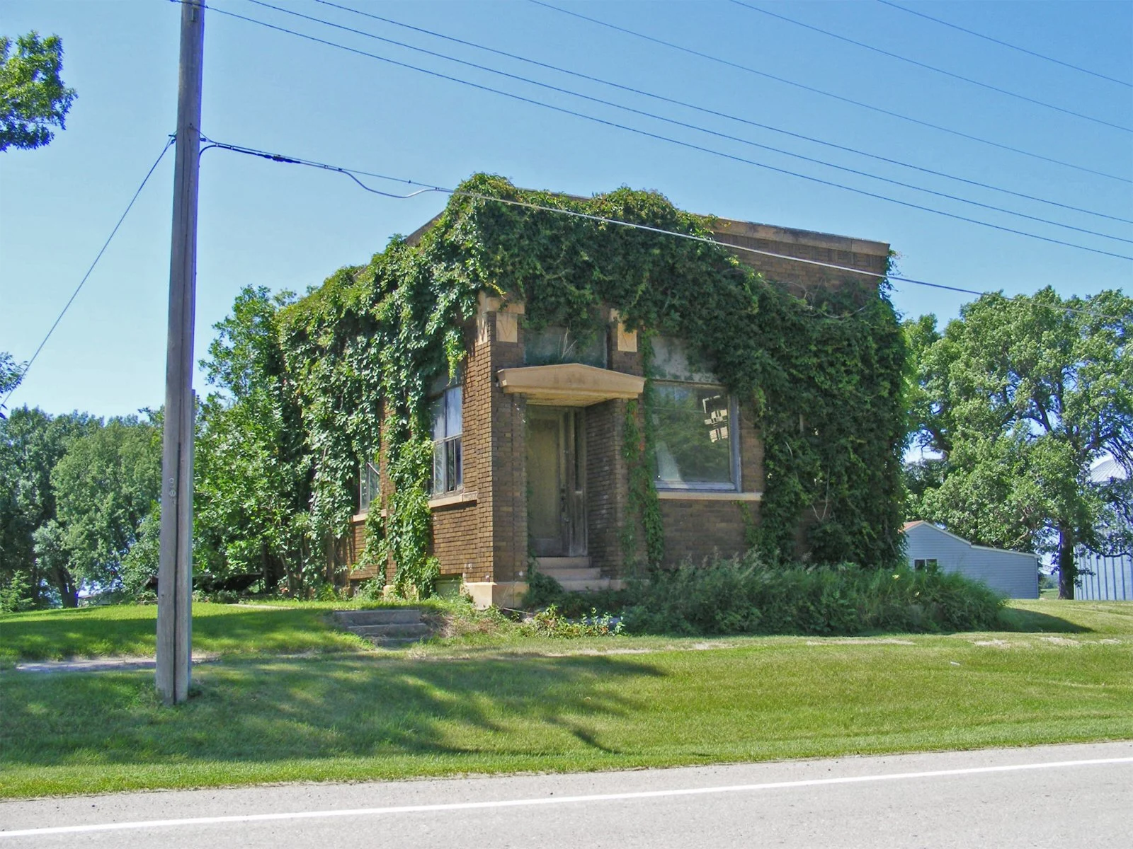 An old brick house covered with green ivy vines and surrounded by green grass and trees, next to a paved road and a utility pole.