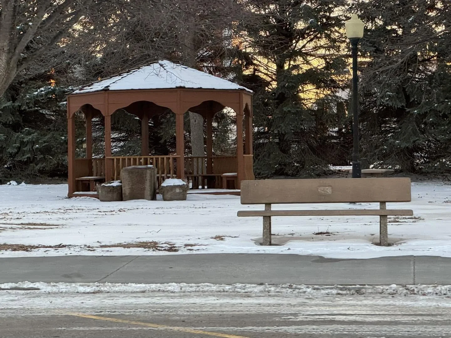 A wooden gazebo with a snow-covered roof in a park, with a bench, street lamp, and snow-covered ground in the foreground, and trees with snow in the background during twilight.