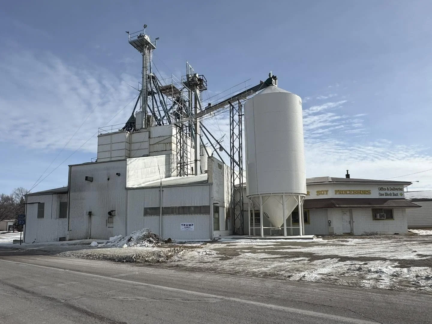 A rural building with grain silos and a small commercial storefront, surrounded by snow and a clear blue sky.