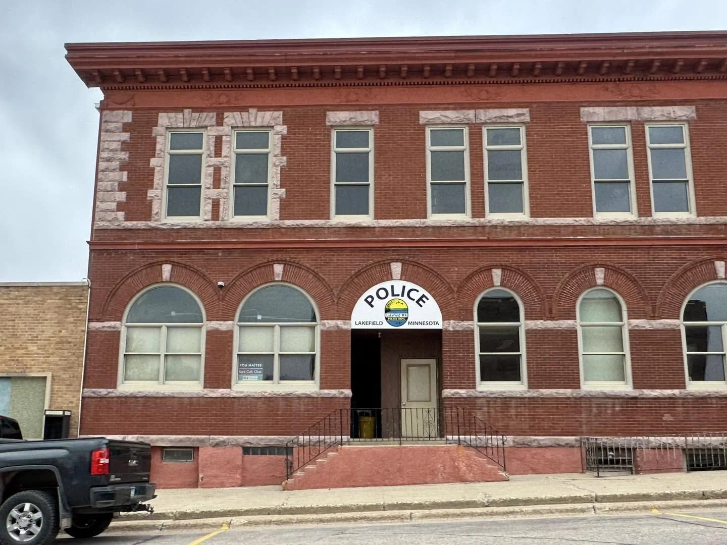 Red brick building with a police station sign, located in Lakefield, Minnesota, featuring arched and rectangular windows, a staircase, and a black truck parked in front.