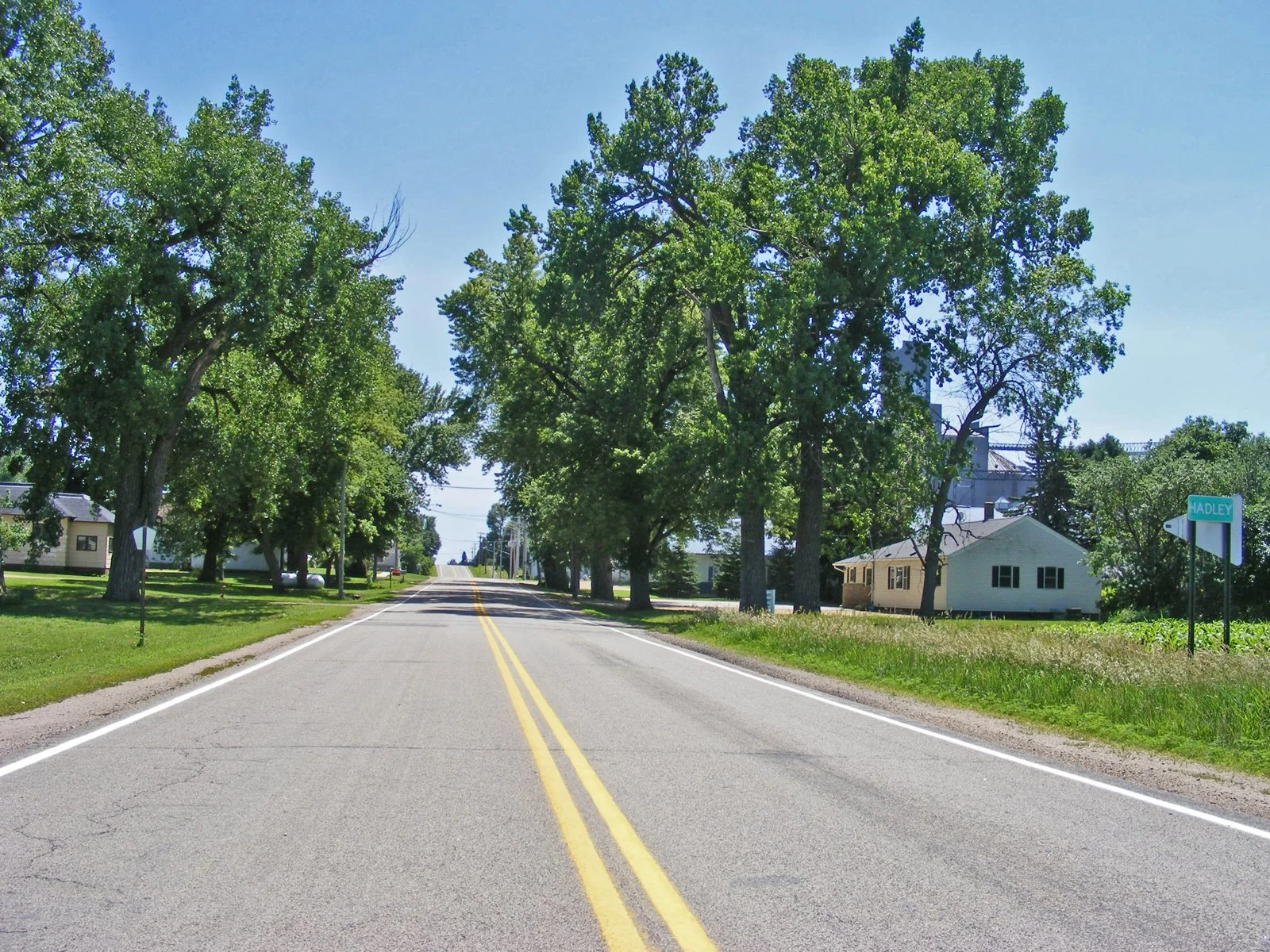A rural street scene with trees lining both sides of the road, houses in the background, a clear blue sky, and a street sign that says 'Jadley' at the corner.