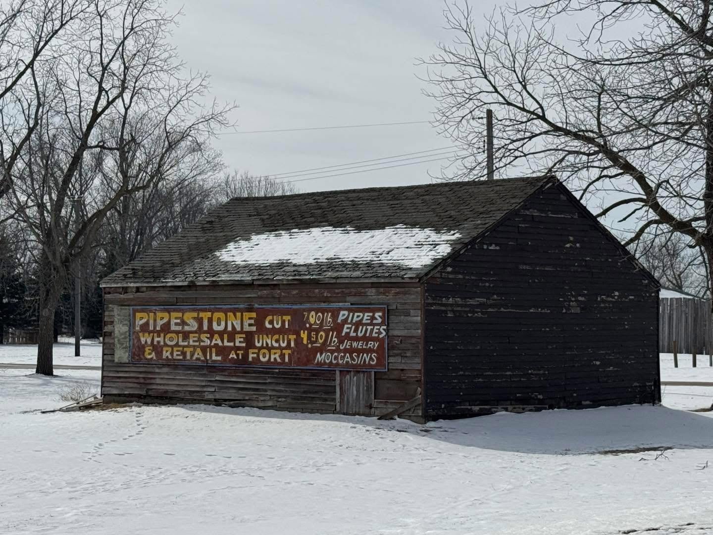 An old wooden building with a snow-covered roof and a fading sign advertising pipestone, wholesale and retail pipes, flutes, jewelry, moccasins, and other items, situated in a snowy landscape with bare trees in the background.