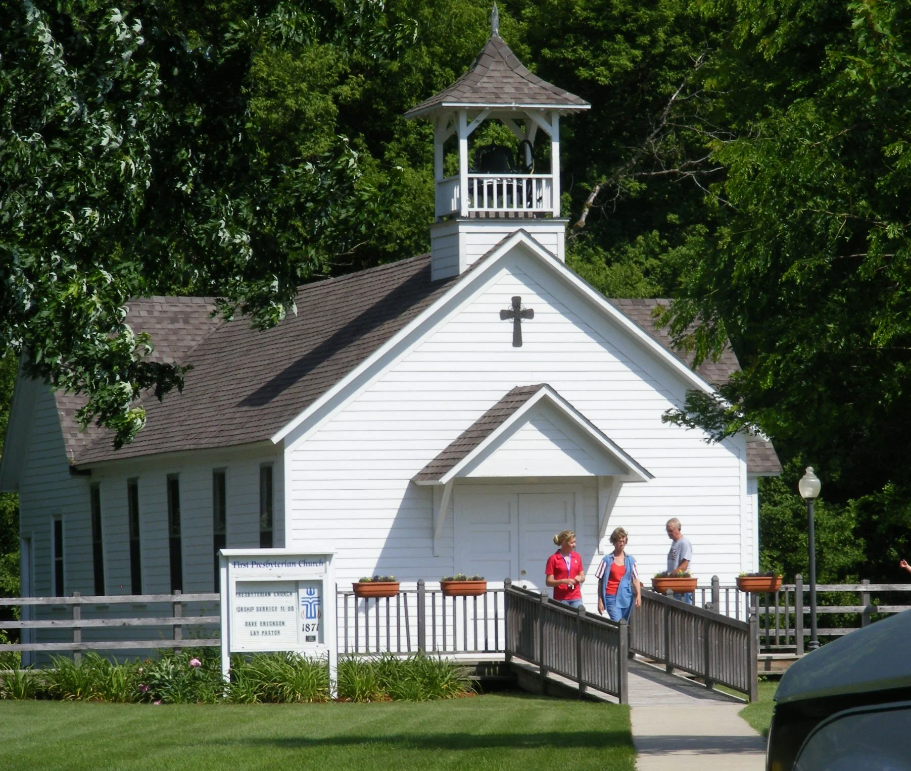 A white Presbyterian church with a small steeple, cross on the front, and a sign in front that reads 'First Presbyterian Church.' There are three people walking near the entrance, surrounded by green trees and a grassy lawn.