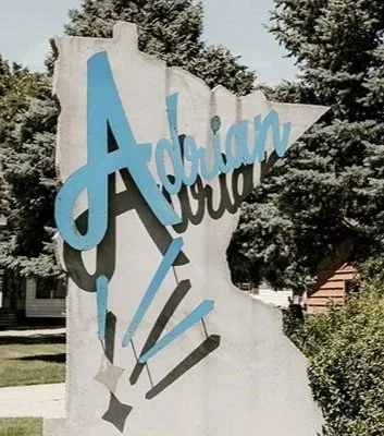 A white rock sign with blue and gray lettering reading 'Adrian, Montana' surrounded by trees and a residential area.