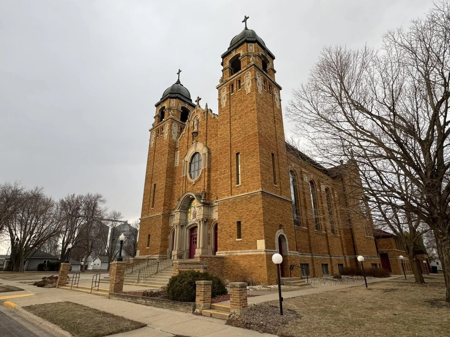 A historic church built with red brick, featuring twin bell towers, arched windows, and a central entrance with steps, situated in a small town with leafless trees and a cloudy sky.
