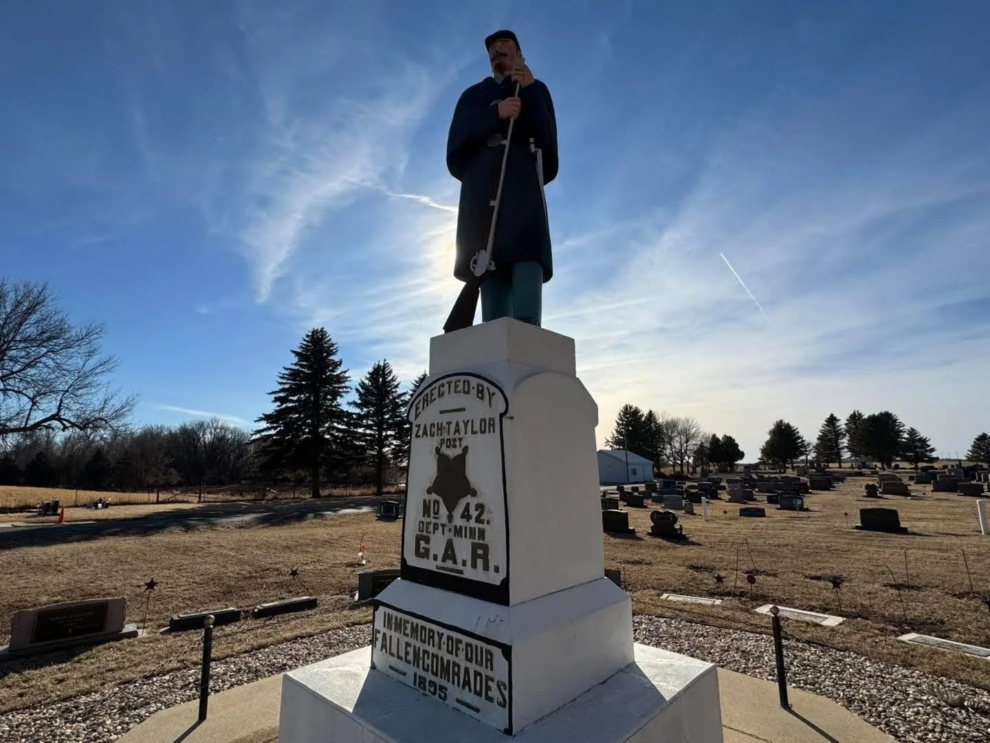 A statue of Abraham Lincoln holding a handrail, standing on a tall white pedestal, in a cemetery with numerous gravestones and trees, under a partly cloudy sky with the sun behind the statue.