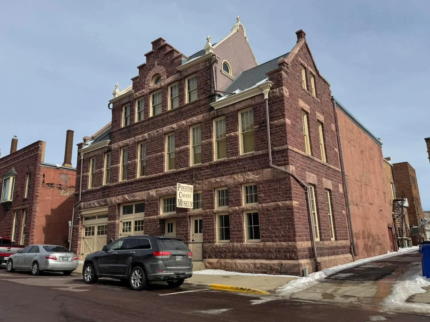 Stone building with a sign that reads 'Pipestone County Museum' in Pipestone, Minnesota, with parked cars in front and melted snow on the ground.