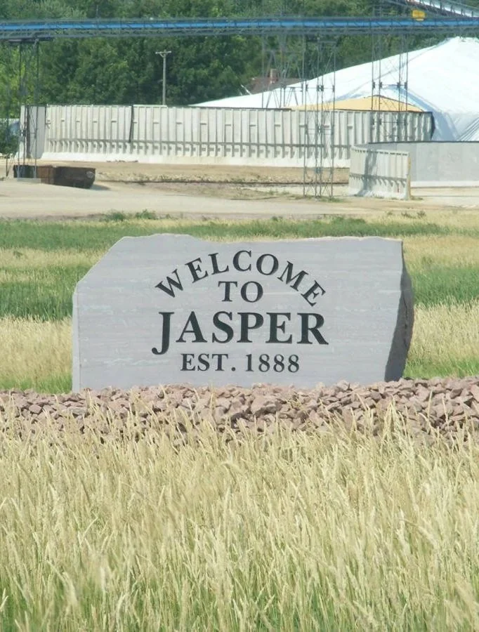 Welcome to Jasper sign in a field with grass and rocks, with industrial structures and trees in the background.